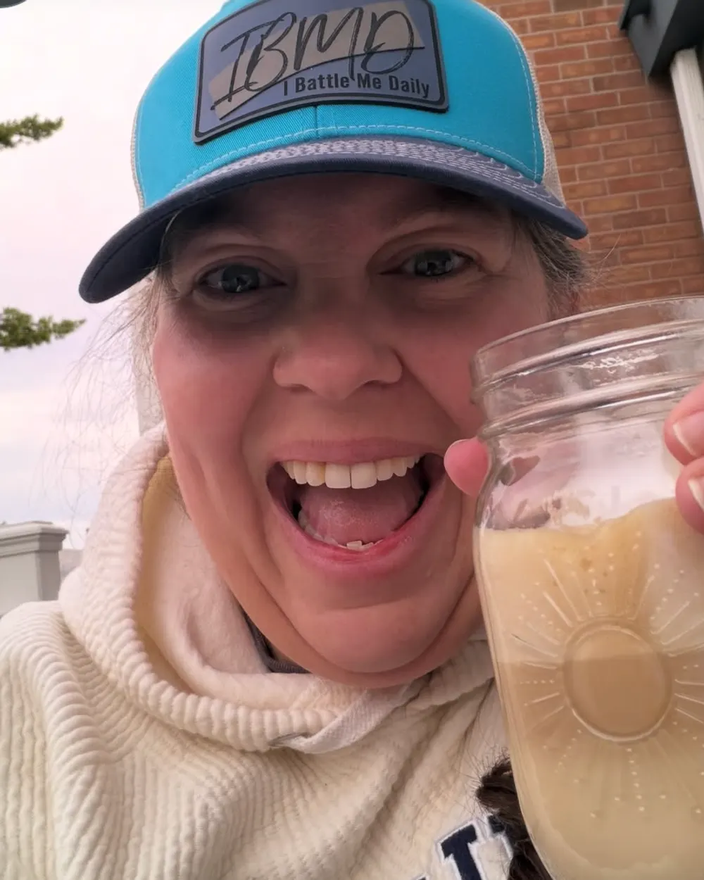 livinginthehotpink smiling in an I Battle Me Daily trucker hat, holding an iced drink outdoors
